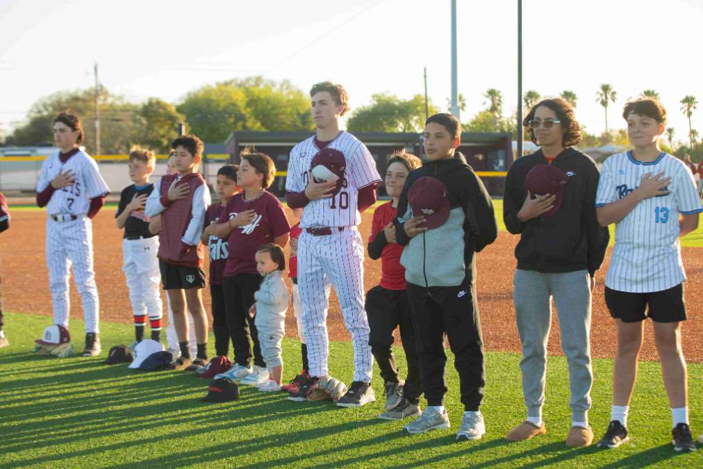 A group of young baseball players and children stand in a line on the field with their right hands over their hearts, some holding baseball caps, during a pre-game ceremony at sunset.