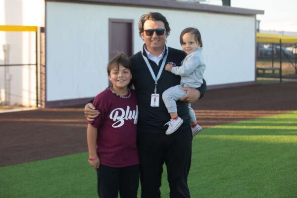 A man stands on a sports field with a young child in his arms and an older child beside him. The older child wears a maroon shirt, while the man and the younger child wear black and gray. All are smiling at the camera.