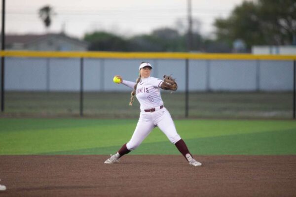 A softball player in a white and maroon uniform prepares to throw the ball on a field, with her arm extended back and glove forward. The outfield fence and trees are visible in the background.