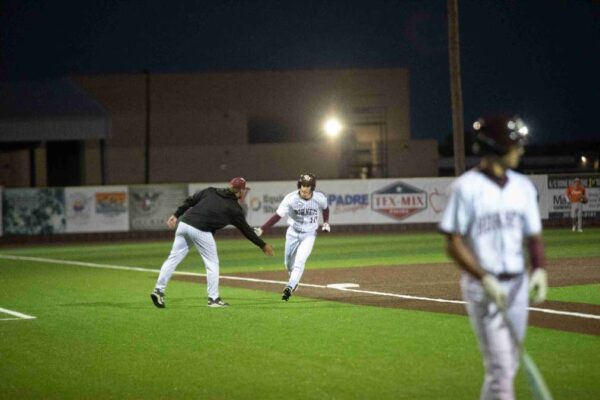 A baseball player in white runs past a coach, who reaches out for a high-five, while another player stands near home plate on a lit field at night.