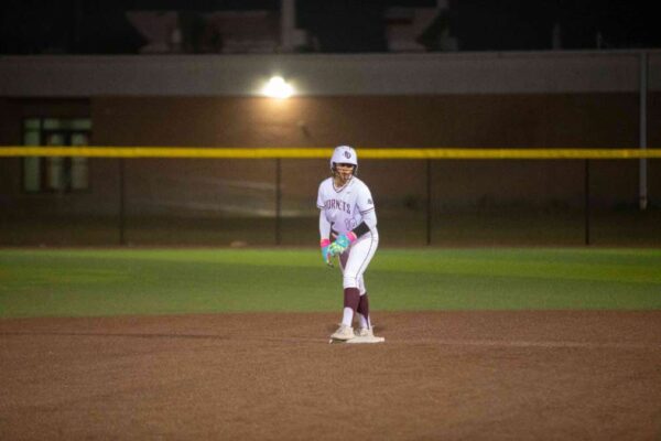 A softball player in a white and maroon uniform stands ready on the field at night, illuminated by field lights, with a yellow outfield fence and a building in the background.