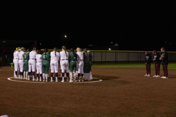 A group of softball players in green and white uniforms stand in a circle on the field at night, heads bowed. A few coaches and players stand off to the side, observing the moment.