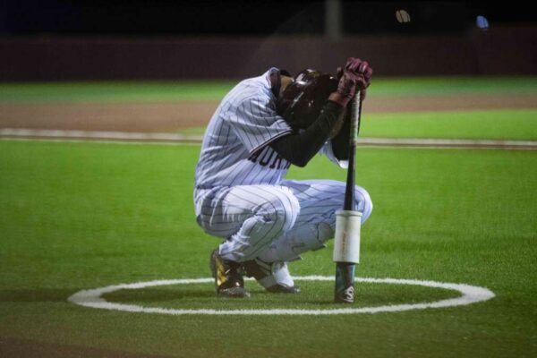 A baseball player in a striped uniform kneels in the on-deck circle, head bowed and resting on his arms atop his bat, appearing deep in thought or prayer on a brightly lit field at night.