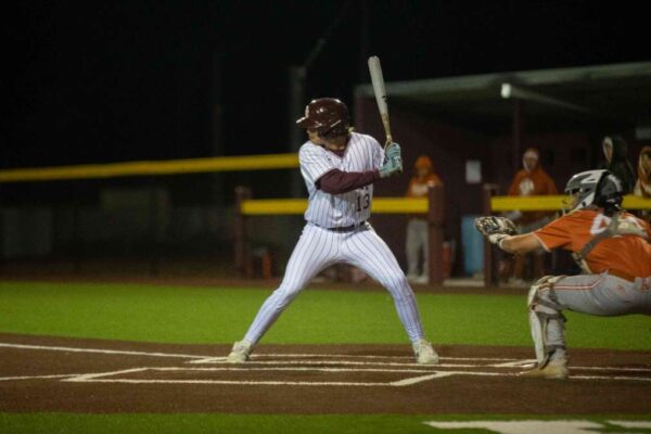 A baseball player in a white and maroon pinstripe uniform stands ready to bat as the catcher and umpire, both in orange gear, prepare behind home plate under stadium lights at night.