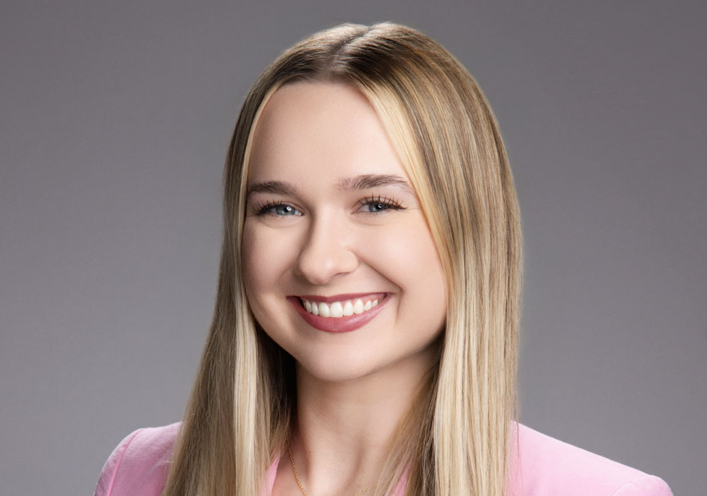 A young woman with long straight blonde hair smiles at the camera. She is wearing a light pink blazer and standing in front of a plain gray background.