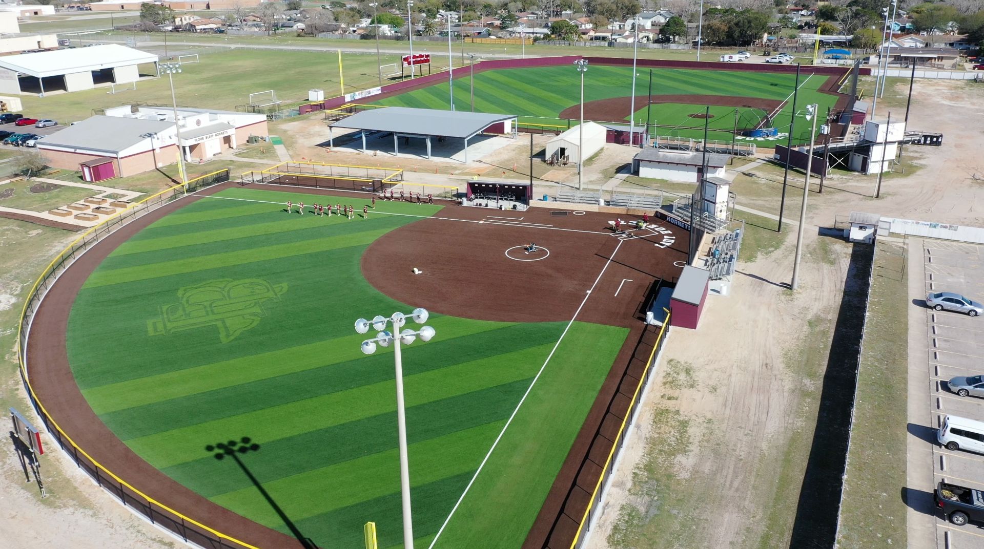 Aerial view of a well-maintained softball field with artificial turf, brown infield, and green outfield. Players stand near the dugout. Nearby are additional sports fields, buildings, and a parking lot.
