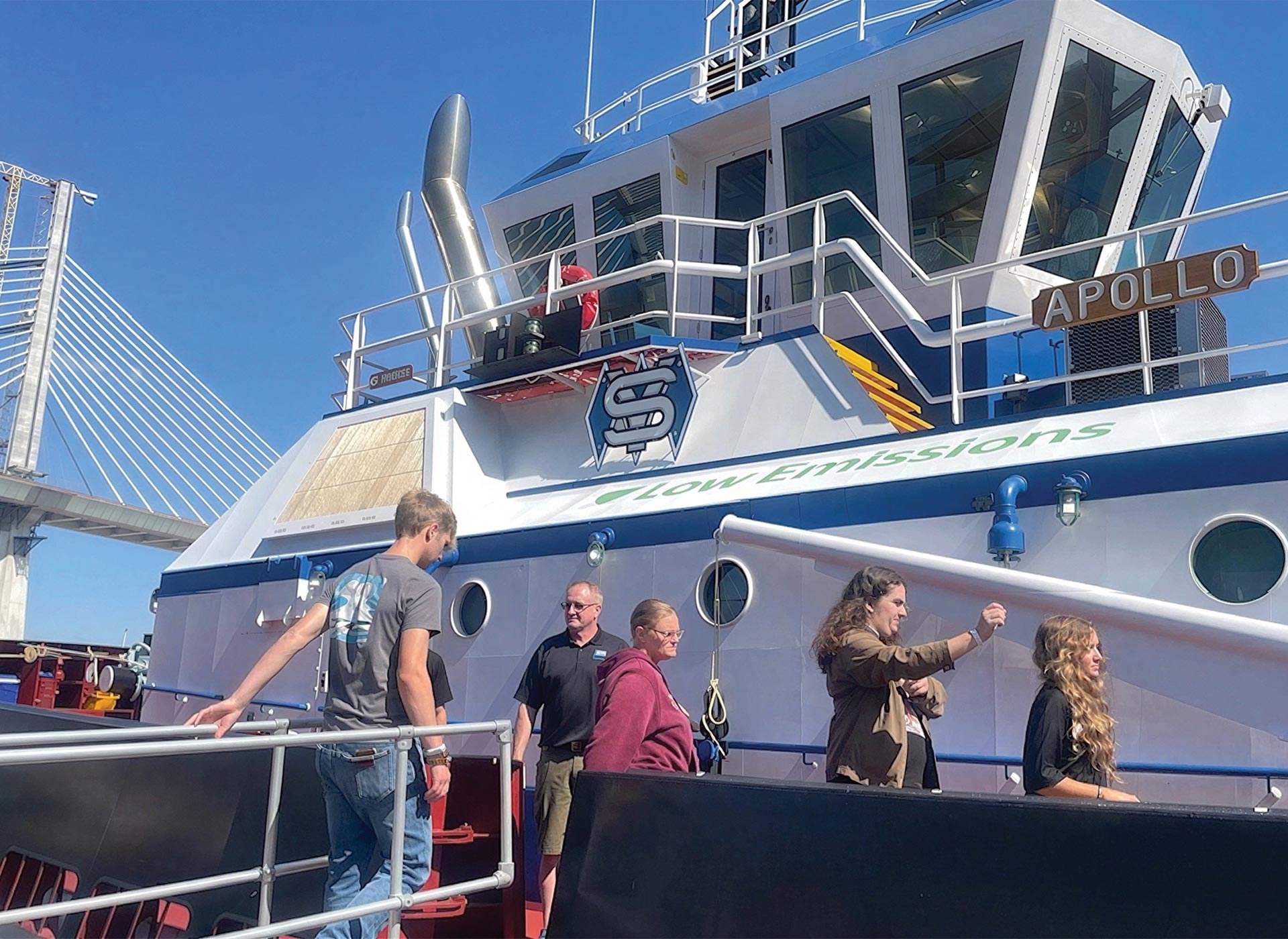 People walk up a gangway to board a white and blue boat named APOLLO with Low Emissions written on the side. A large bridge and clear blue sky are visible in the background.