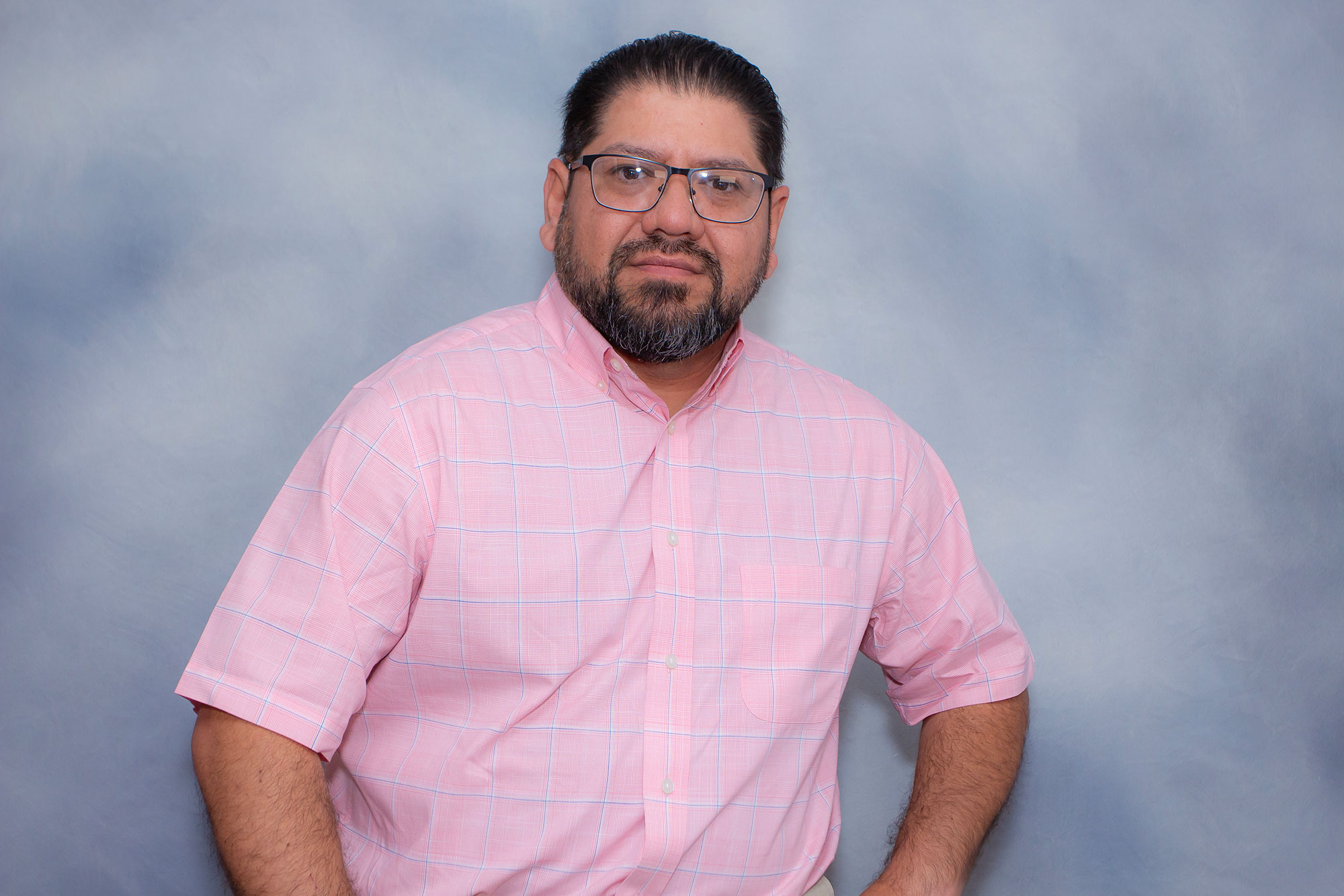 Man with dark hair, beard, and glasses wearing a light pink plaid short-sleeve shirt, posing in front of a light blue-gray studio backdrop with a neutral expression.