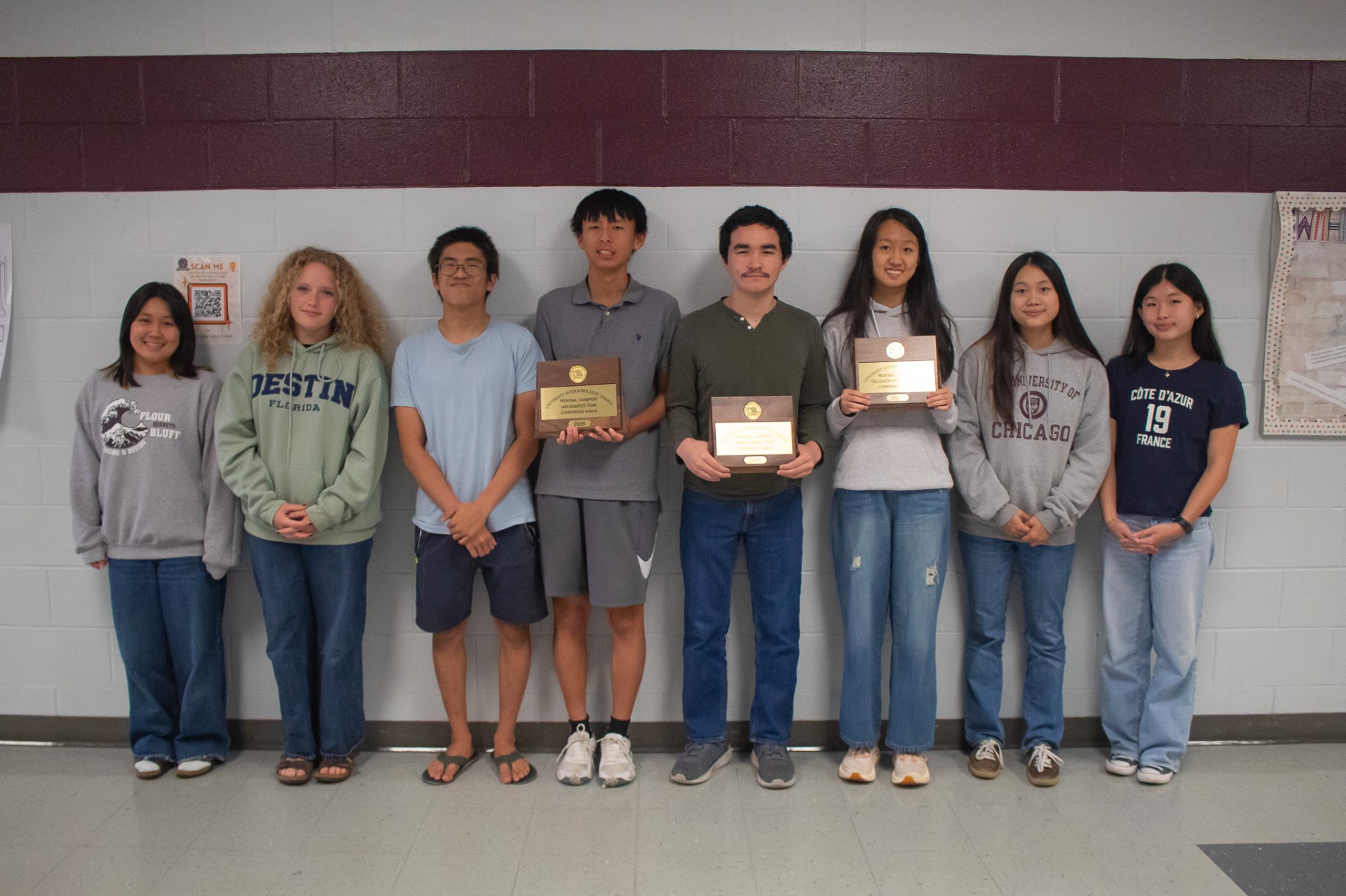 Eight students stand in a row indoors against a maroon and gray wall. Three students in the center hold plaques and awards, while the others stand with hands at their sides, all smiling at the camera.