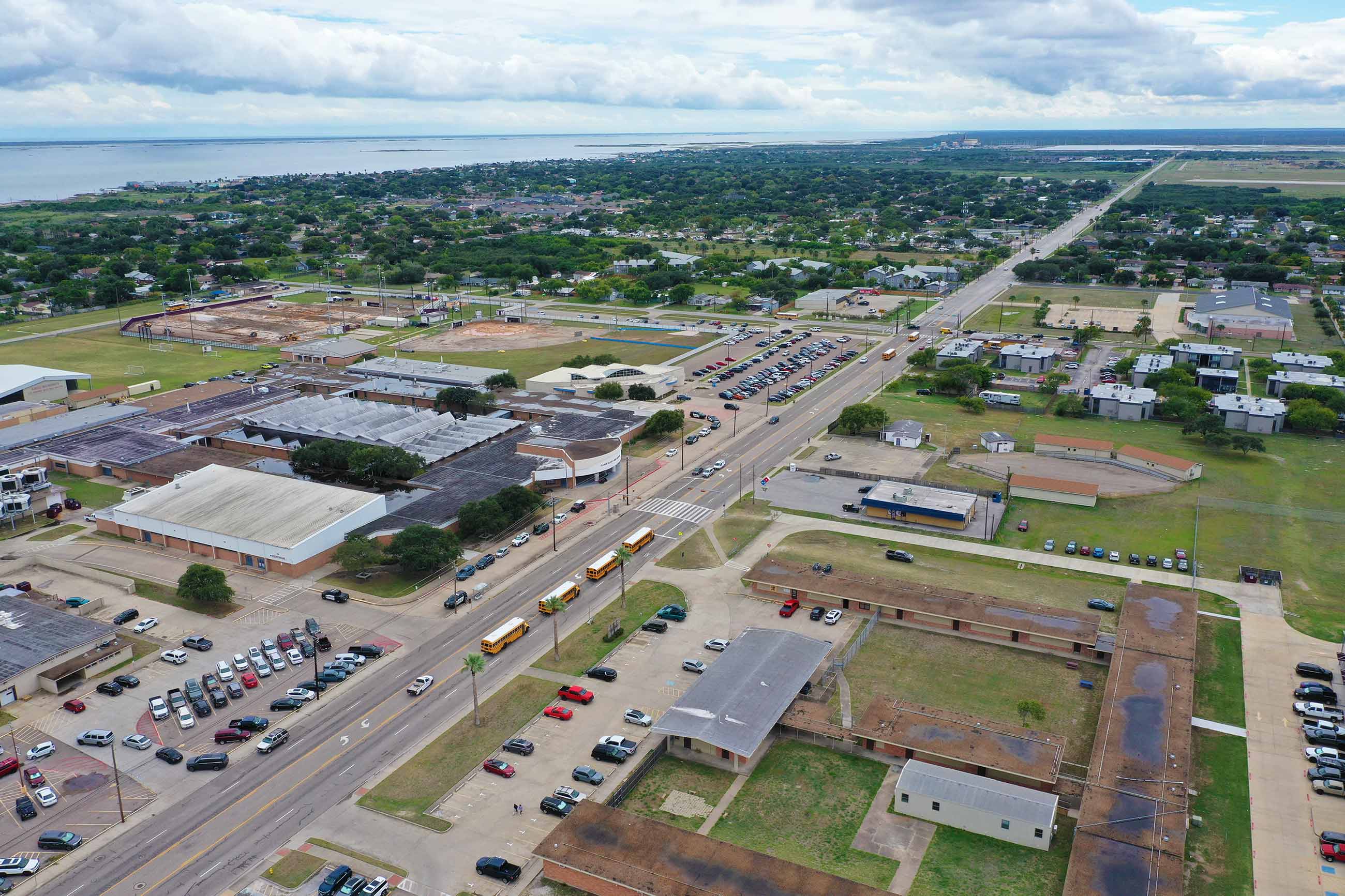 Aerial view of a suburban town with a wide road, parking lots, school buildings, scattered houses, and green spaces under a cloudy sky; a body of water is visible in the distance.