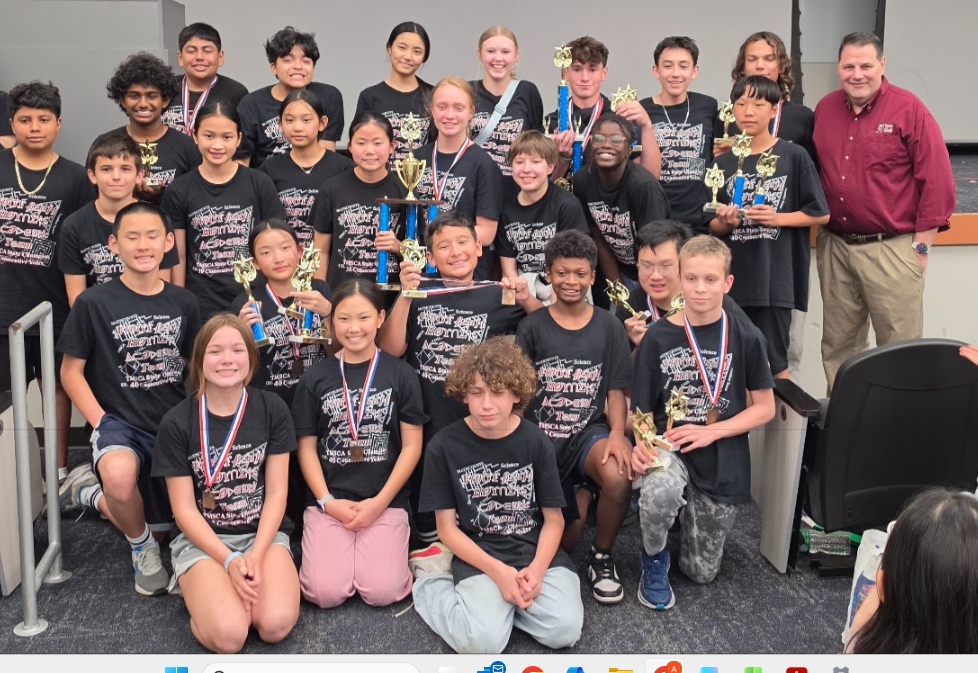 A group of students wearing matching black shirts with medals and trophies pose together indoors, smiling at the camera with an adult standing on the right.