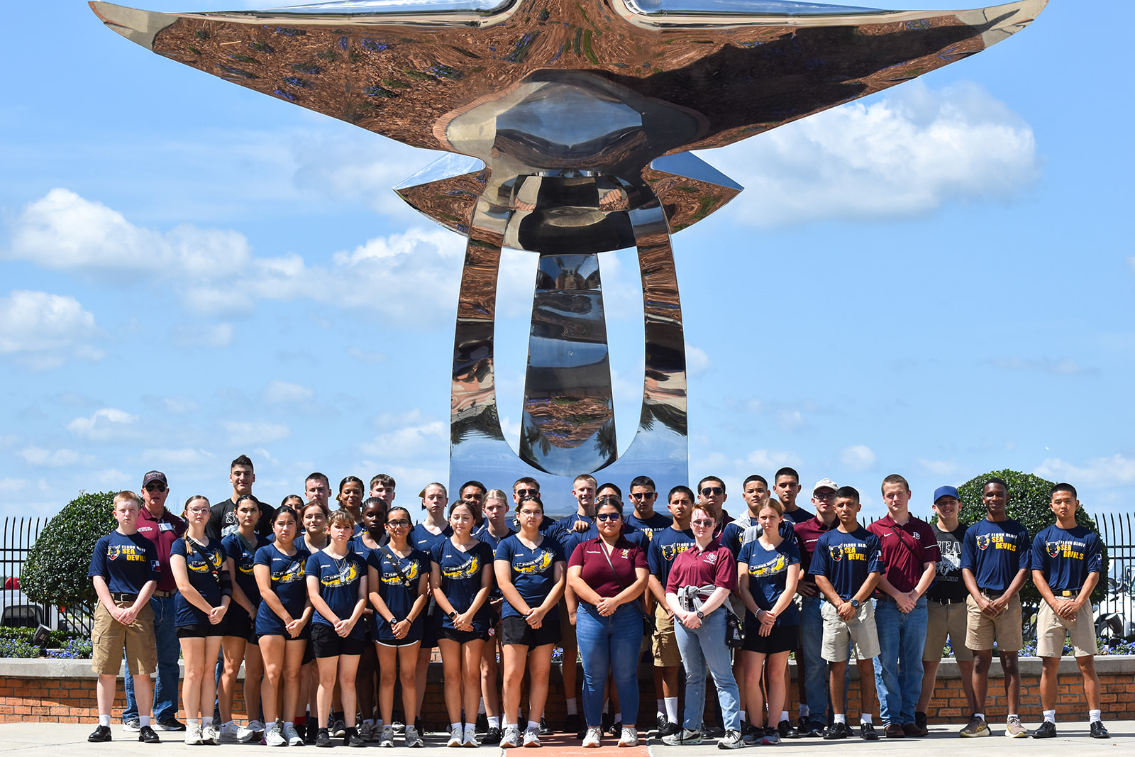 Grand Nationals 2026 A group of young adults and teens pose in front of a large, shiny, abstract metal sculpture outdoors on a sunny day, with blue skies and clouds in the background. Many wear matching navy and yellow shirts.