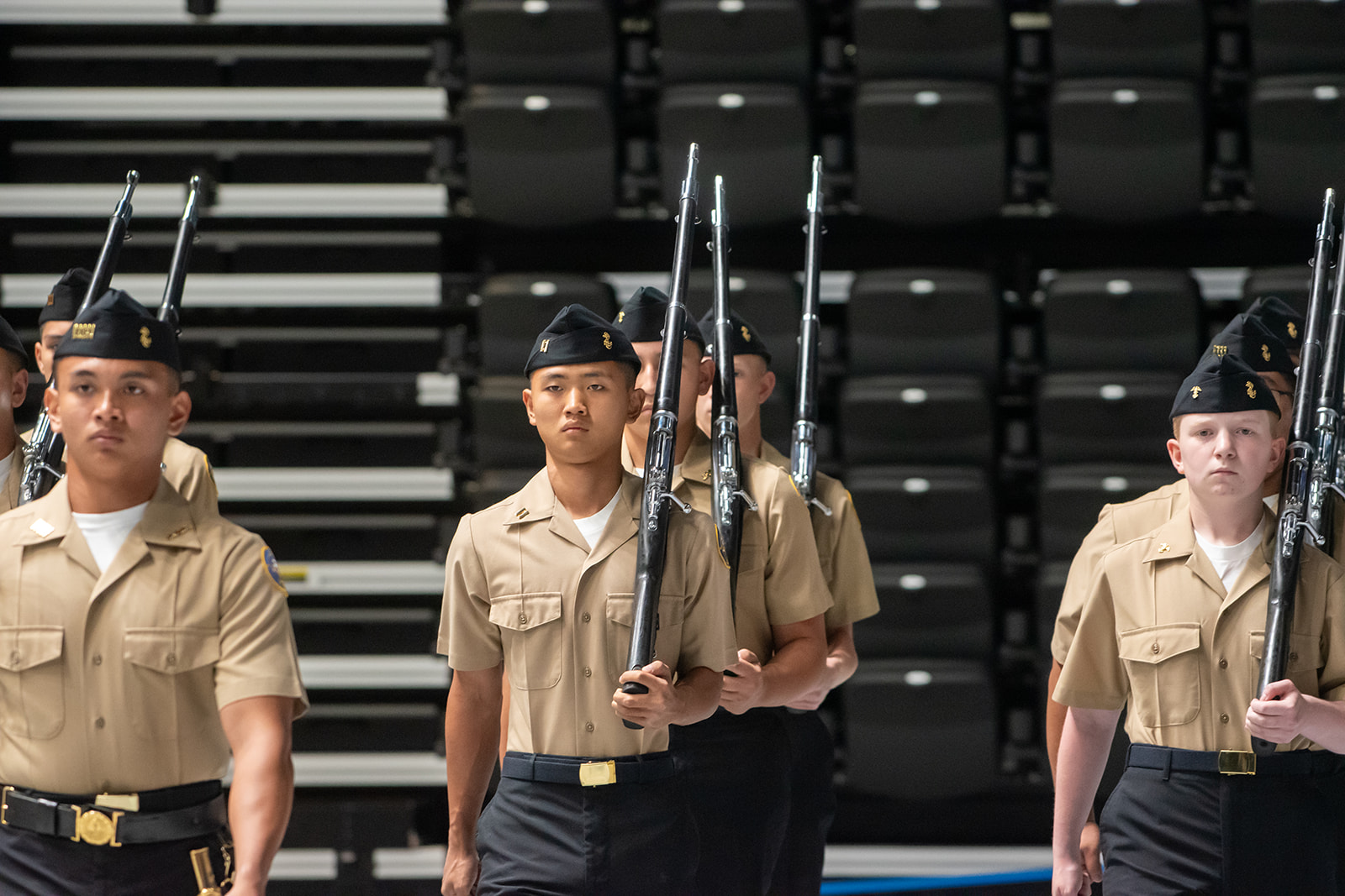 Grand Nationals 2026 A group of uniformed cadets in formation march indoors, holding rifles upright on their shoulders. The background shows empty rows of stadium-style seating.