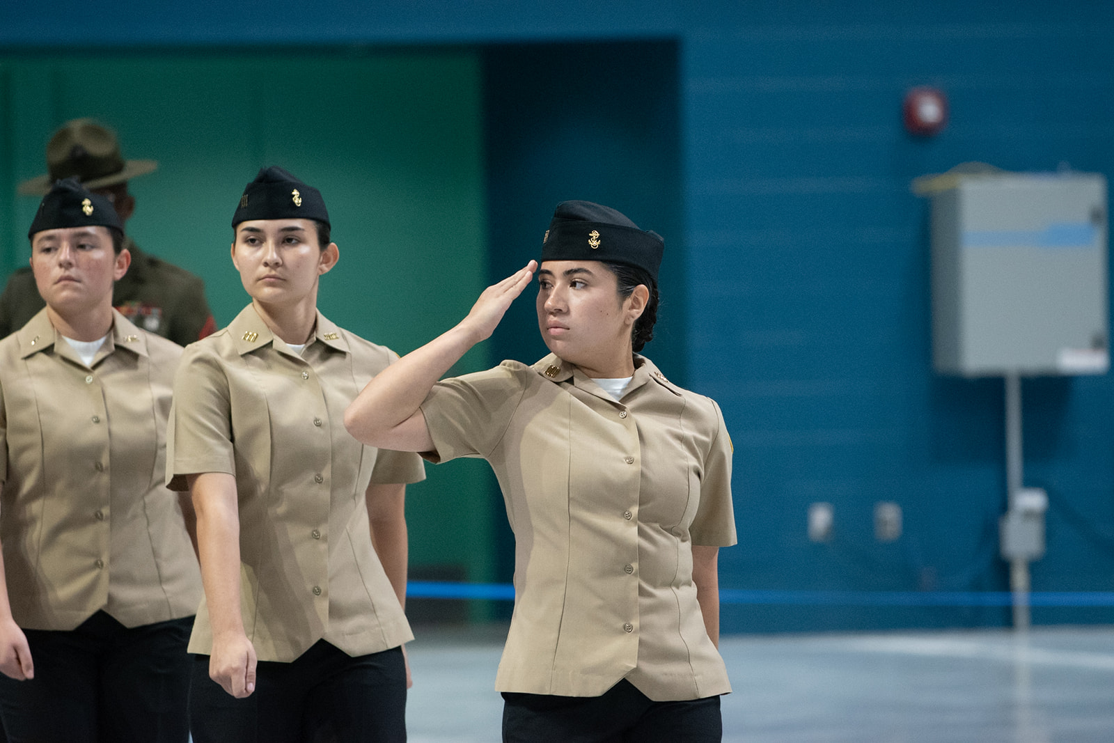 Grand Nationals 2026 Three uniformed women march indoors in formation, with the woman in the center foreground saluting. They are dressed in khaki shirts and dark hats, participating in a military or ROTC event.