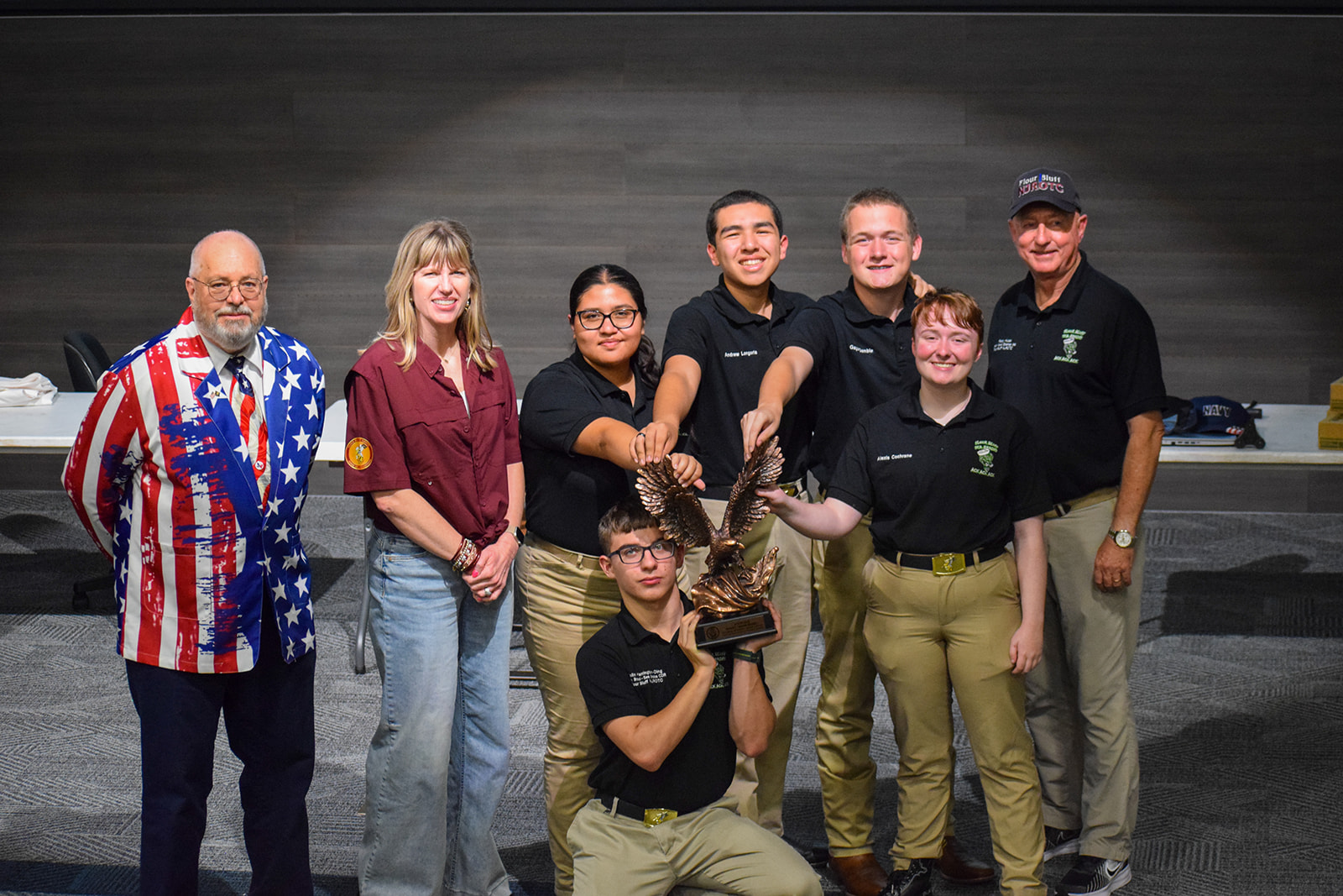 Grand Nationals 2026 A group of eight people, some in matching uniforms, pose indoors. Five in the center hold up a trophy with eagle sculptures. Two people in casual shirts and one in an American flag blazer stand beside them, smiling.