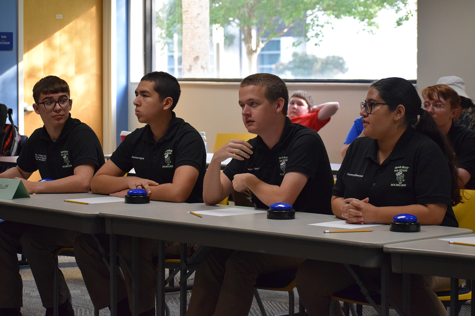 Grand Nationals 2026 Four students wearing matching black shirts sit at a table with buzzers and pencils, appearing focused and ready to participate in a quiz or academic competition in a classroom setting.