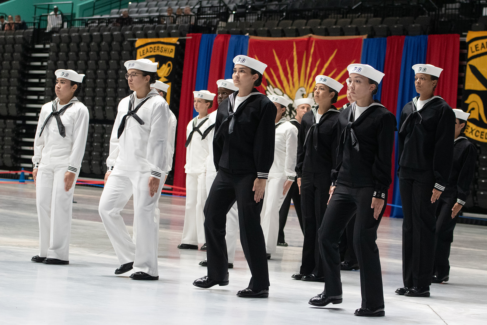 Grand Nationals 2026 A group of Navy recruits in white and black uniforms stand in formation indoors during a ceremony, with colorful banners and empty seating in the background.
