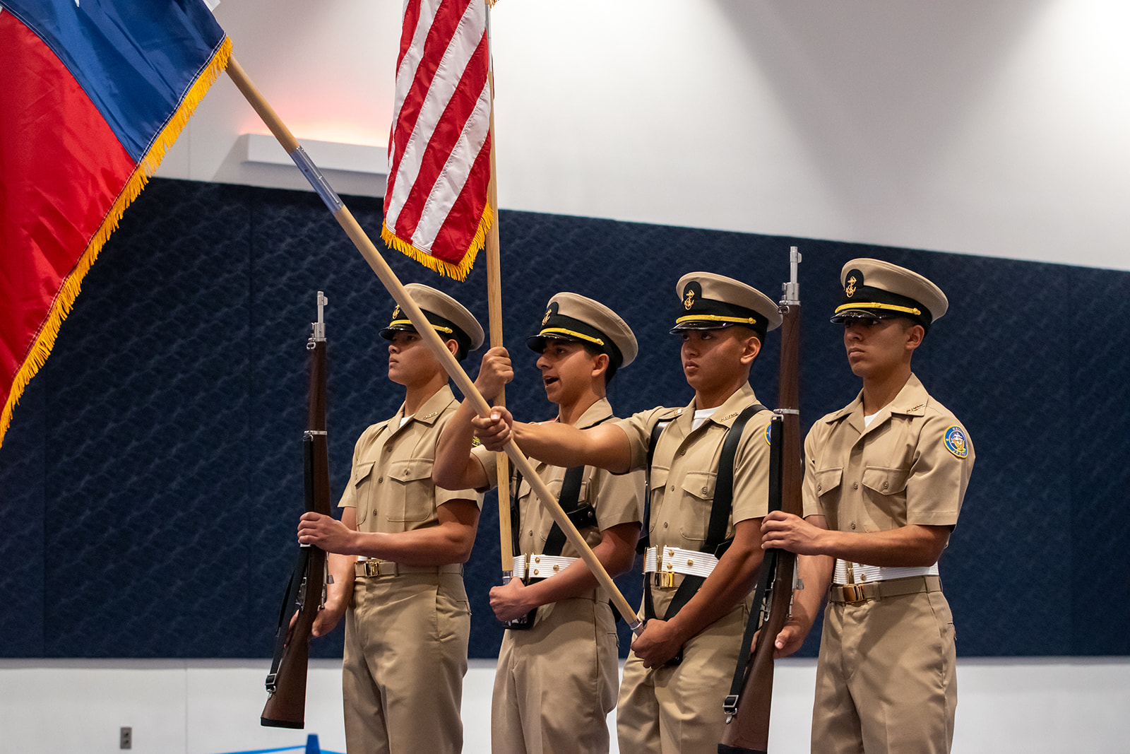 Four uniformed cadets stand in a row, two holding rifles while the middle two hold flags, including the American flag, during a formal ceremony indoors.
