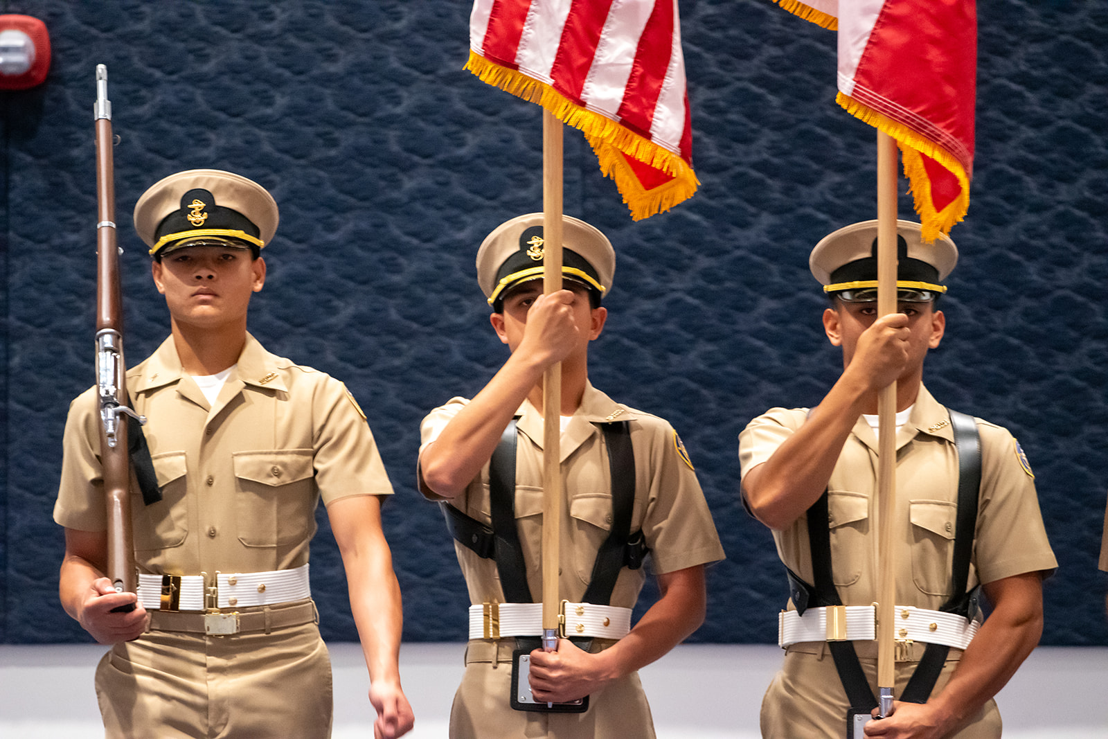 Grand Nationals 2026 Three uniformed service members stand in formation indoors. The two on the right hold flags, while the one on the left holds a rifle. They wear khaki uniforms with dark ties and peaked caps.