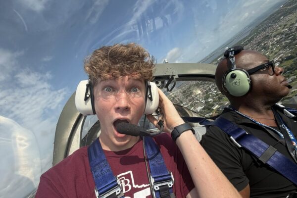 Hornet Aviation First Flight 1 A young man with curly hair looks surprised while wearing headphones and sitting in an airplane cockpit beside another person, who is calmly looking out the window. Both are wearing seatbelts, and the sky is visible outside.