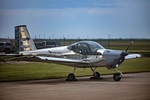 Hornet Aviation First Flight (10) (1) A small, silver, single-engine airplane with tricycle landing gear is parked on a concrete surface near a grassy field, under a cloudy sky.