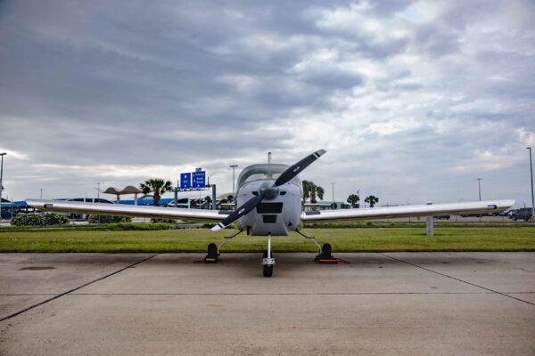 Hornet Aviation First Flight (11) (1) A small single-engine propeller airplane is parked on a concrete surface under a cloudy sky, with grassy areas, signs, and palm trees in the background.