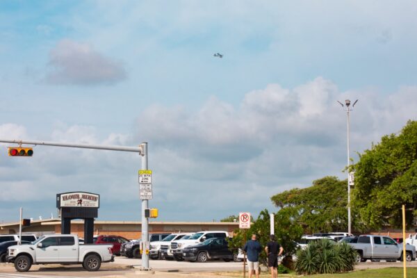 Hornet Aviation First Flight 3 A busy street corner with parked cars, a traffic light, and two people standing near greenery. The sign reads Blackbeards Restaurant. An airplane is visible flying in the cloudy sky above.