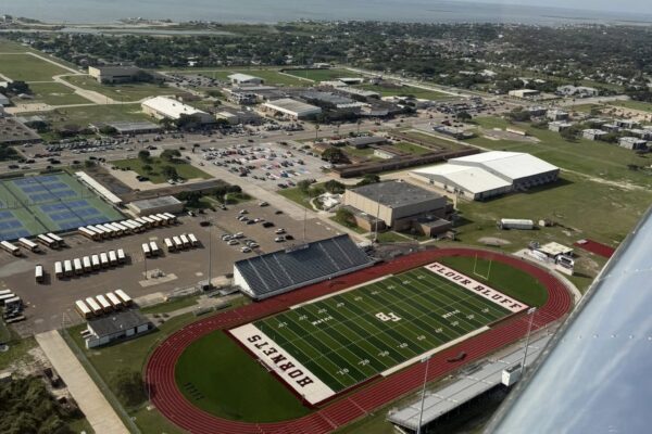 Hornet Aviation First Flight 4 Aerial view of a high school football stadium with a red track labeled “BOBCATS,” surrounded by school buildings, tennis courts, parking lots, and a coastal town visible in the background.