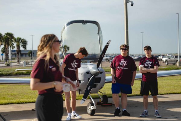 Hornet Aviation First Flight (6) (1) Four young adults in matching maroon shirts stand near a small white airplane on a sunny day, with one woman in the foreground and three men near the aircraft. Palm trees and a building are visible in the background.