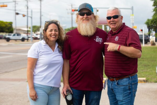 Hornet Aviation First Flight 6 Three adults pose and smile outdoors on a sidewalk. One woman wears a white polo, and two men wear maroon shirts; one man has a long beard and sunglasses. There are cars, utility poles, and traffic lights in the background.