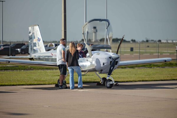 Hornet Aviation First Flight (7) (1) Four people stand next to a small, single-engine airplane with an open cockpit at an airport, having a conversation on the tarmac near a runway. The sky is clear and other cars are visible in the background.