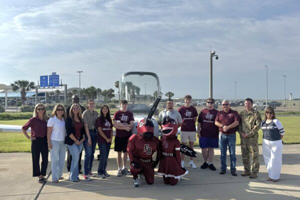 Hornet Aviation First Flight 7 A group of people, including mascots and someone in military uniform, stand and kneel together outdoors on a sunny day, posing for a photo on a concrete surface with grass and blue sky in the background.