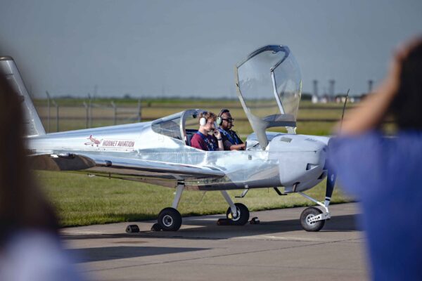 Hornet Aviation First Flight (8) (1) Two people sit inside a small, silver single-engine airplane with the cockpit canopy open, parked on an airport runway. The airport field and distant buildings are visible in the background.