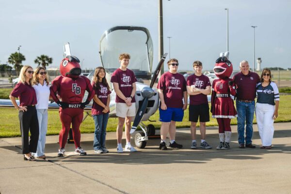Hornet Aviation First Flight (9) (1) A group of people, including two mascots in maroon costumes and several individuals in maroon shirts, stand in front of a small aircraft on a sunny day.