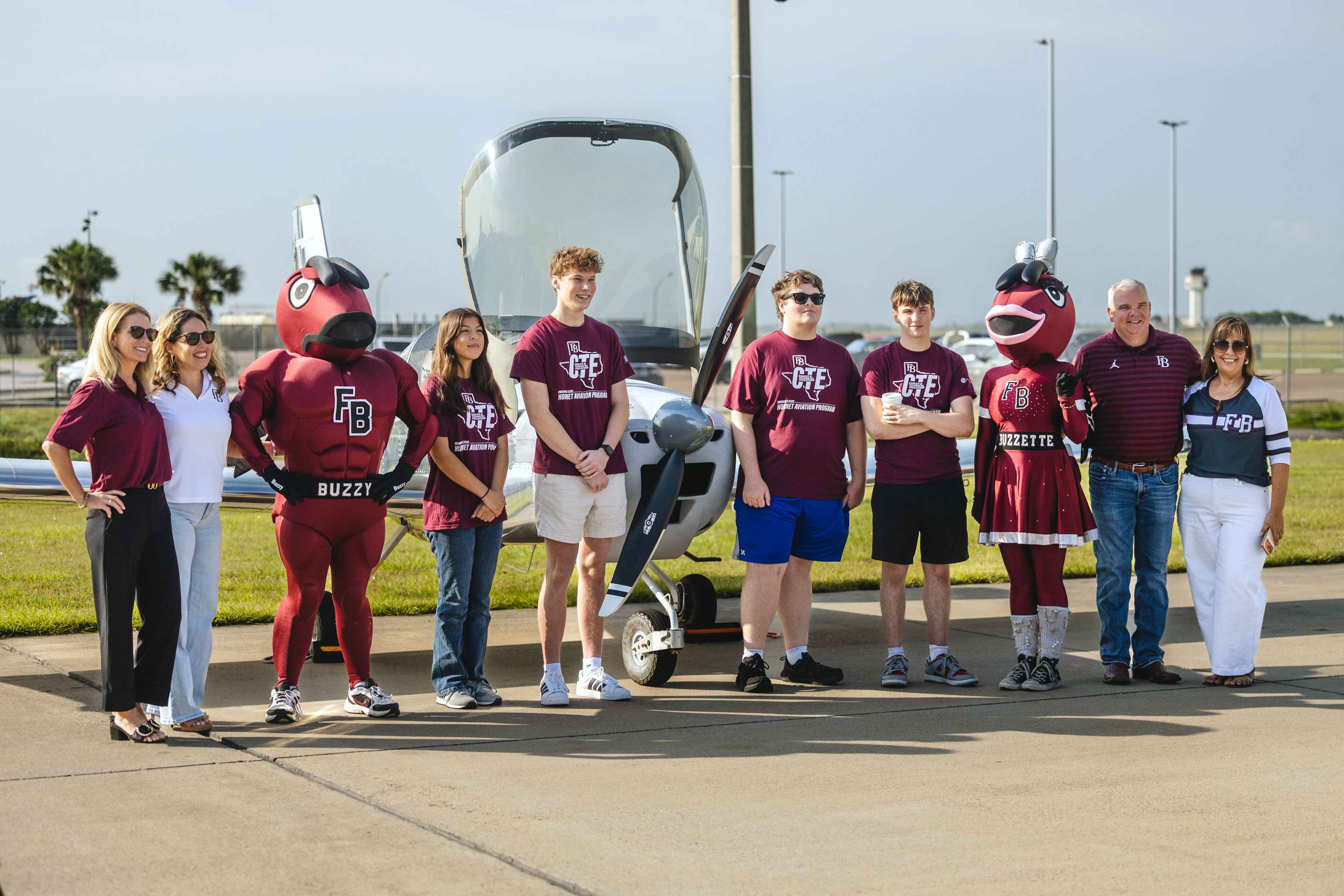 A group of people, including two mascots in maroon costumes and several individuals in maroon shirts, stand in front of a small aircraft on a sunny day.