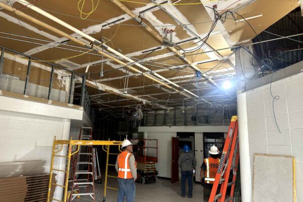 JH Renovation 5 Several construction workers wearing safety vests and helmets stand in a room with exposed ceiling beams, pipes, and wires. Ladders and scaffolding are present, indicating ongoing renovation work.
