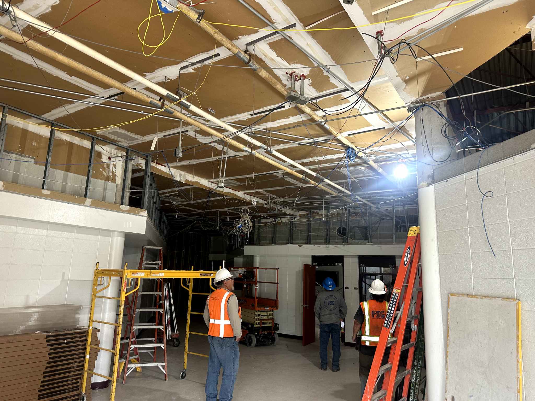 Several construction workers wearing safety vests and helmets stand in a room with exposed ceiling beams, pipes, and wires. Ladders and scaffolding are present, indicating ongoing renovation work.