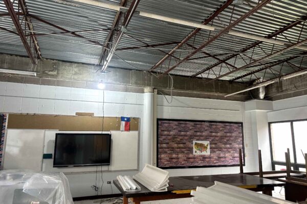JH Renovation 7 A classroom under renovation with exposed ceiling beams, scattered construction materials, a TV mounted on a wall, and a map of the United States on a brick-patterned bulletin board.