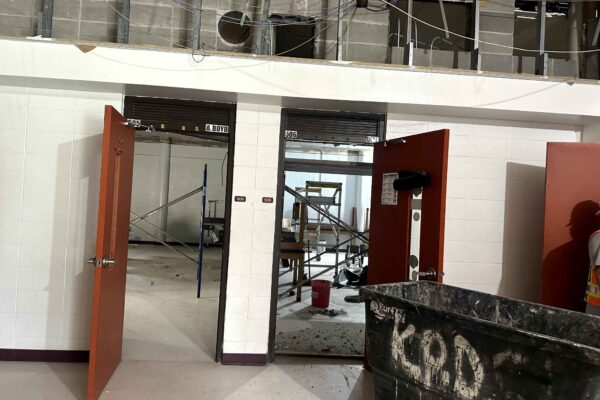 JH Renovation 8 Three open red doors lead to rooms under construction, with exposed ceiling, scaffolding, tools, and debris visible. A worker in a safety vest stands near a large black bin marked “ZOD.”.