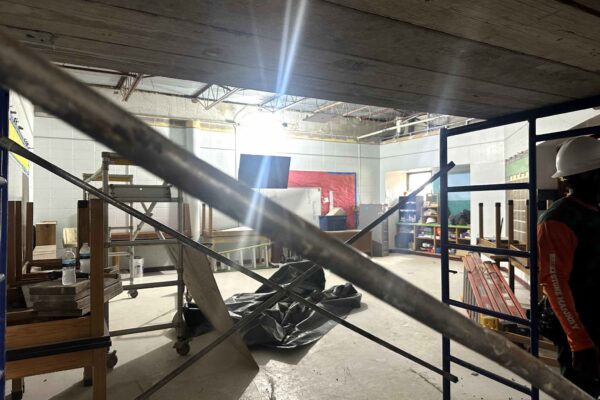 JH Renovation 3 A construction worker wearing a hard hat stands in a cluttered, partially finished room with exposed ceiling beams, scattered building materials, and crossed metal scaffolding in the foreground. Bright light shines from the ceiling.
