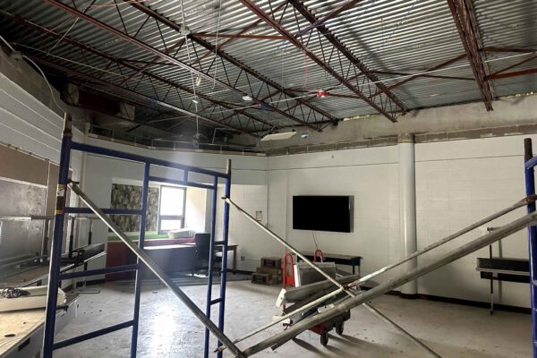 JH Renovation 1 A partially renovated room with exposed ceiling beams, scaffolding, construction materials, and unfinished walls. There are tables, a wall-mounted TV, boxes, and light coming from a window in the back.