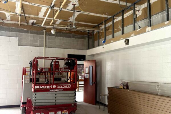 JH Renovation 4 A construction lift is parked inside a partially renovated room with exposed ceiling pipes, unfinished walls, and a wooden slat counter along one side. An open door leads to another area.