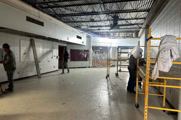 JH Renovation 9 Two workers are renovating a large room with white tiled walls and a partially exposed ceiling. Scaffolding and tools are visible, and tarps cover parts of the space. The room appears empty and under construction.