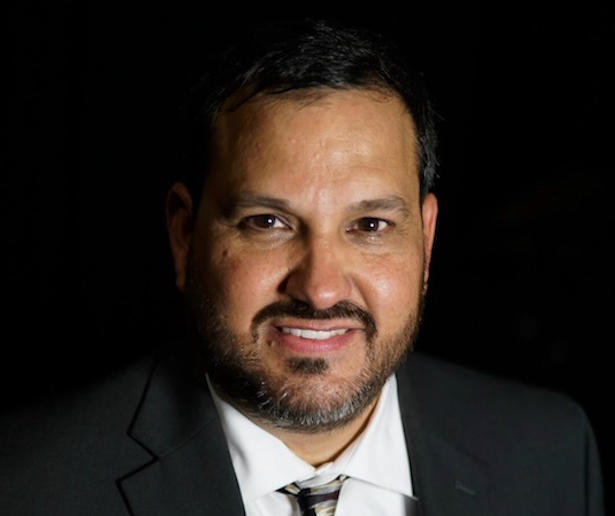 A man with dark hair and a beard, wearing a dark suit, white shirt, and striped tie, smiles against a black background.