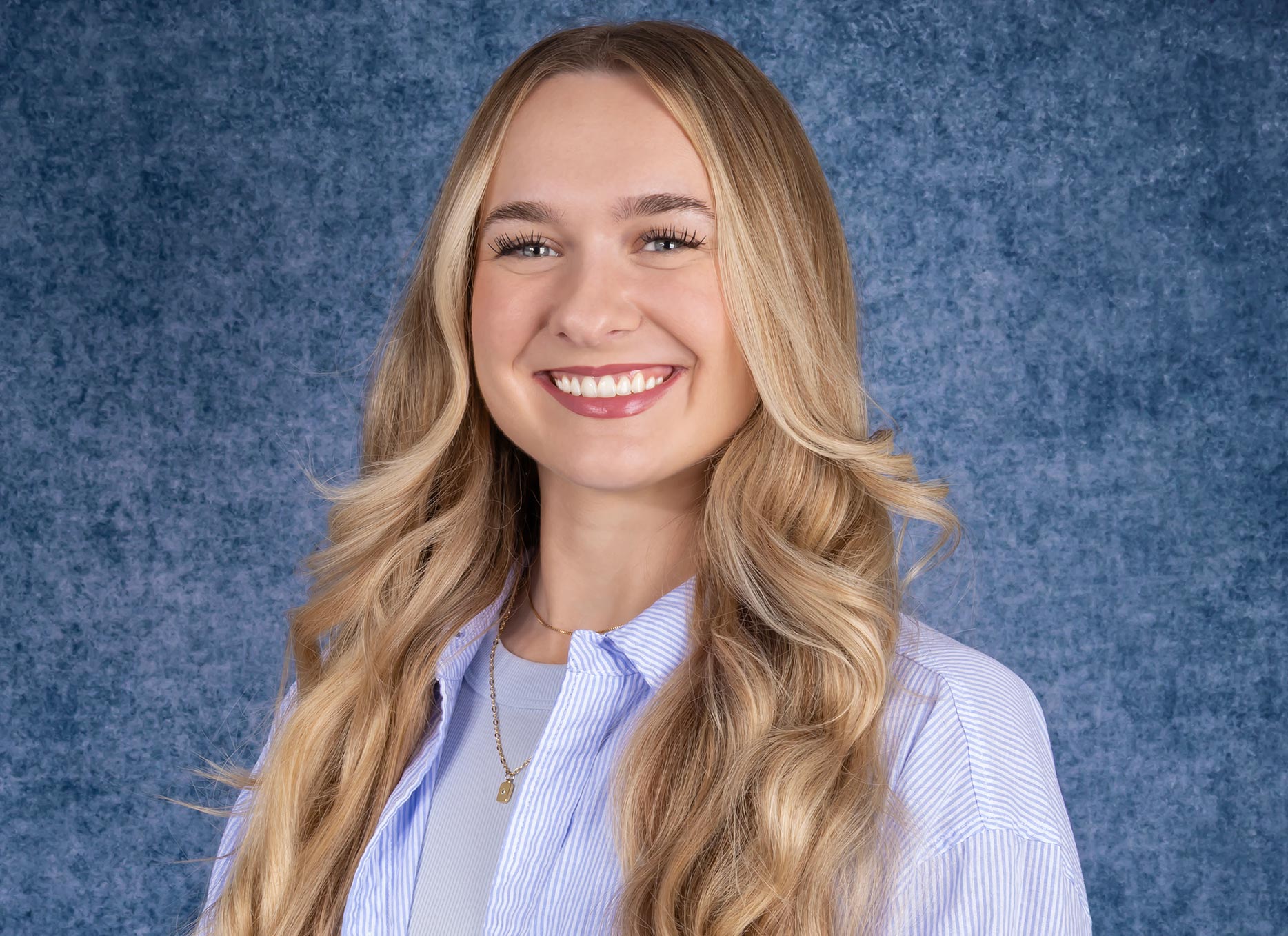 A young woman with long, wavy blonde hair smiles in front of a textured blue background. She is wearing a light blue striped shirt over a light top and a delicate necklace.