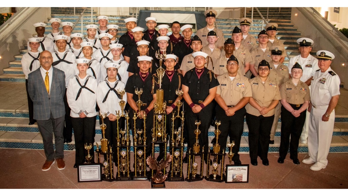 NJROTC National Champions 2026 A large group of naval cadets and officers in uniform pose on stairs, standing behind numerous trophies and plaques. One man in a suit stands at the left end. The scene is well-lit and celebratory.