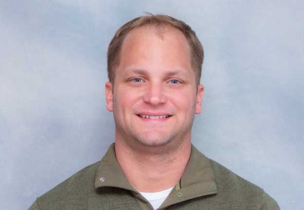 A man with short blond hair is smiling at the camera. He is wearing a light green collared shirt under a green sweater. The background is a plain light grayish blue.