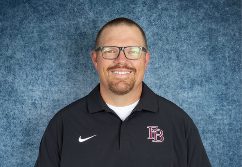 A smiling man with short hair, a goatee, and glasses wears a black collared shirt with a white Nike logo and FB letters on the chest. He stands in front of a textured blue background.