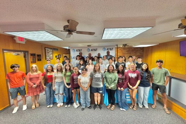 A large group of students and adults pose together indoors in front of an AT&T banner. They are smiling and standing in rows in a brightly lit room with yellow walls and a gray carpet.