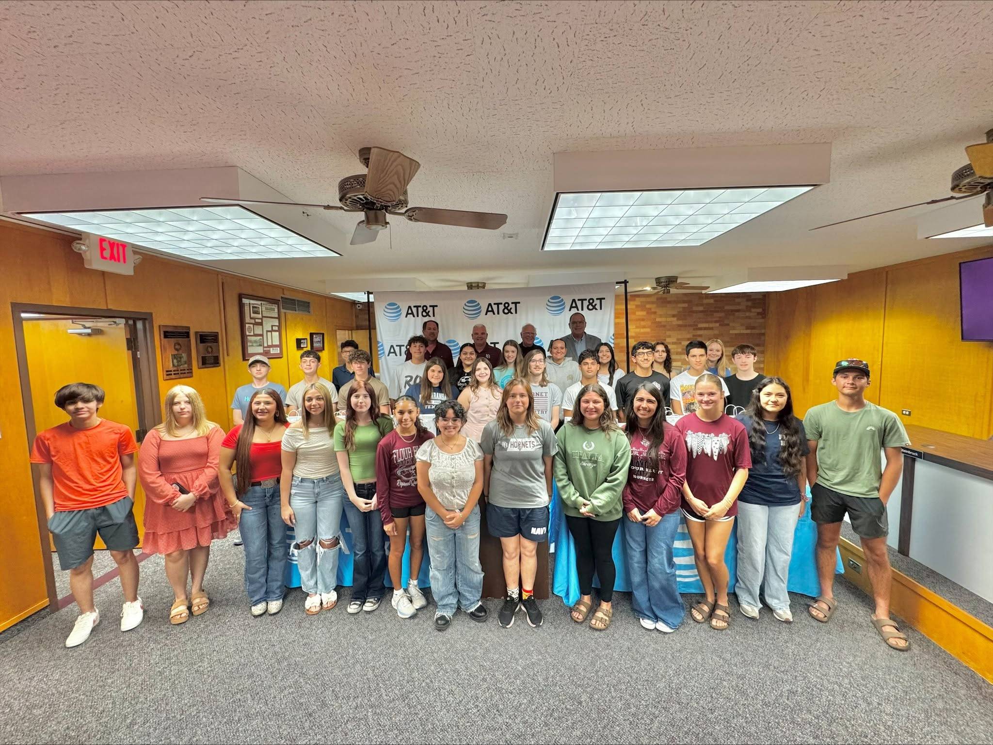 A large group of students and adults pose together indoors in front of an AT&T banner. They are smiling and standing in rows in a brightly lit room with yellow walls and a gray carpet.